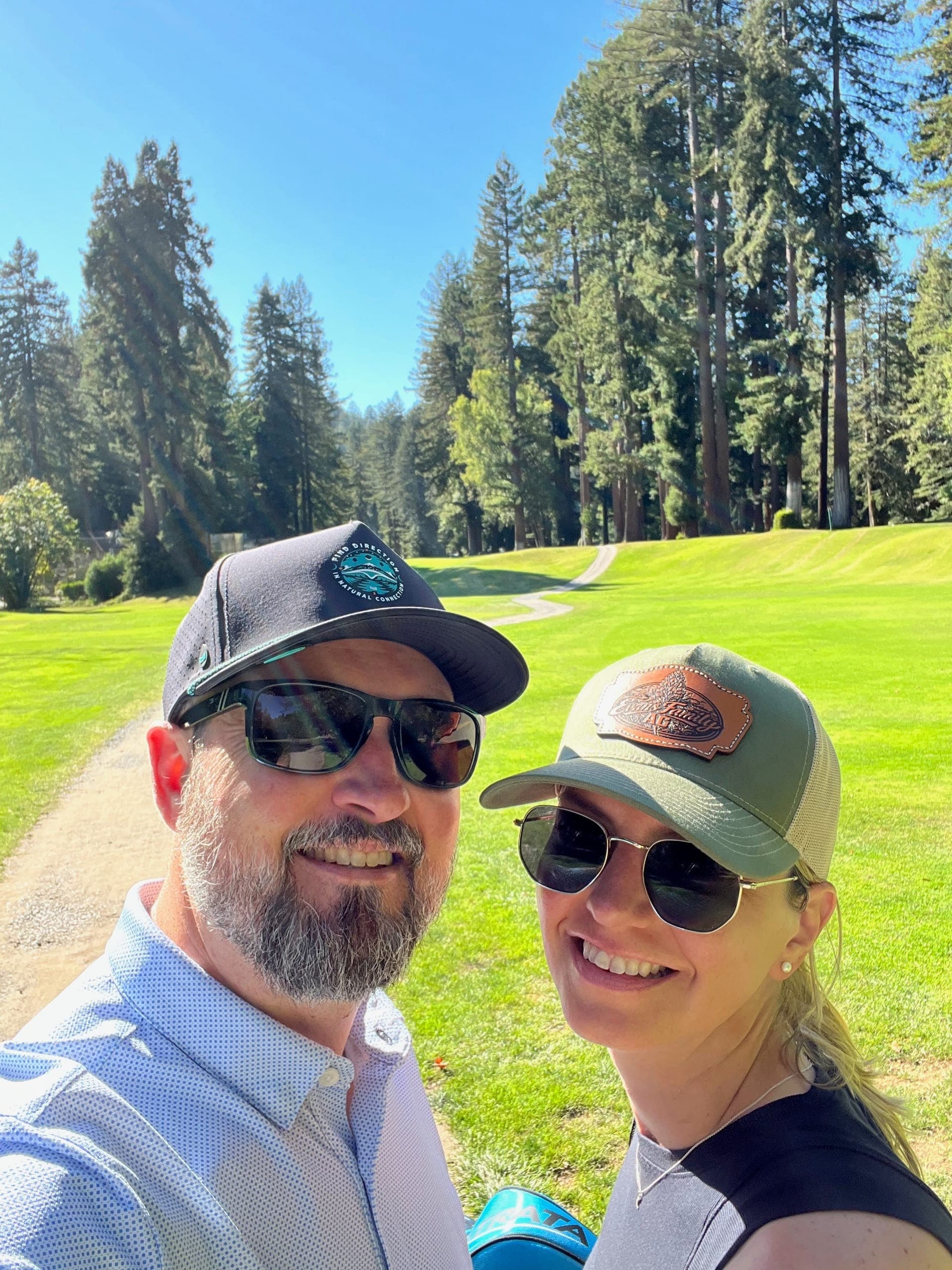 Erik and Sarah taking a selfie in front of a fairway that is outlined in stately redwoods.