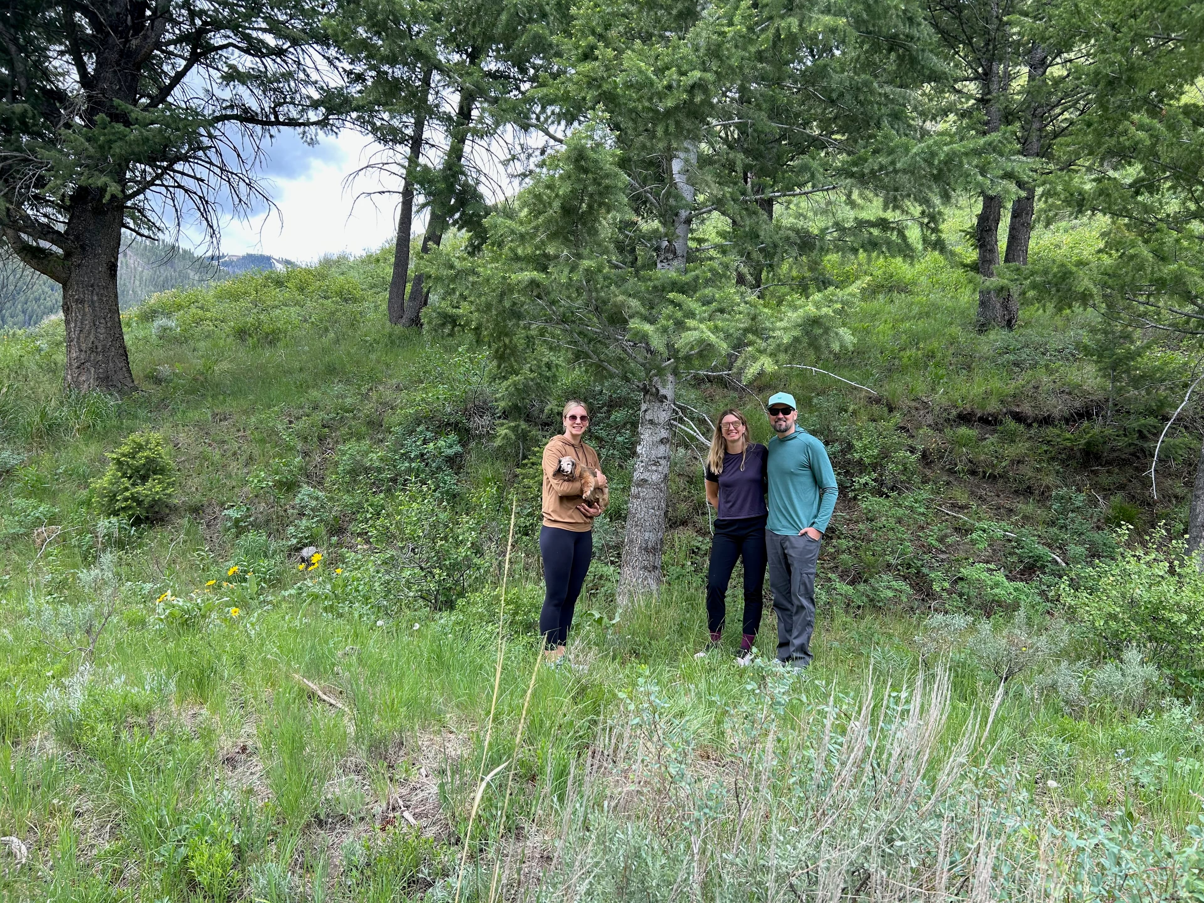 Group photo of Erik, Sarah, and Kayti holding Baby Dog in front of a tree on a green hillside.