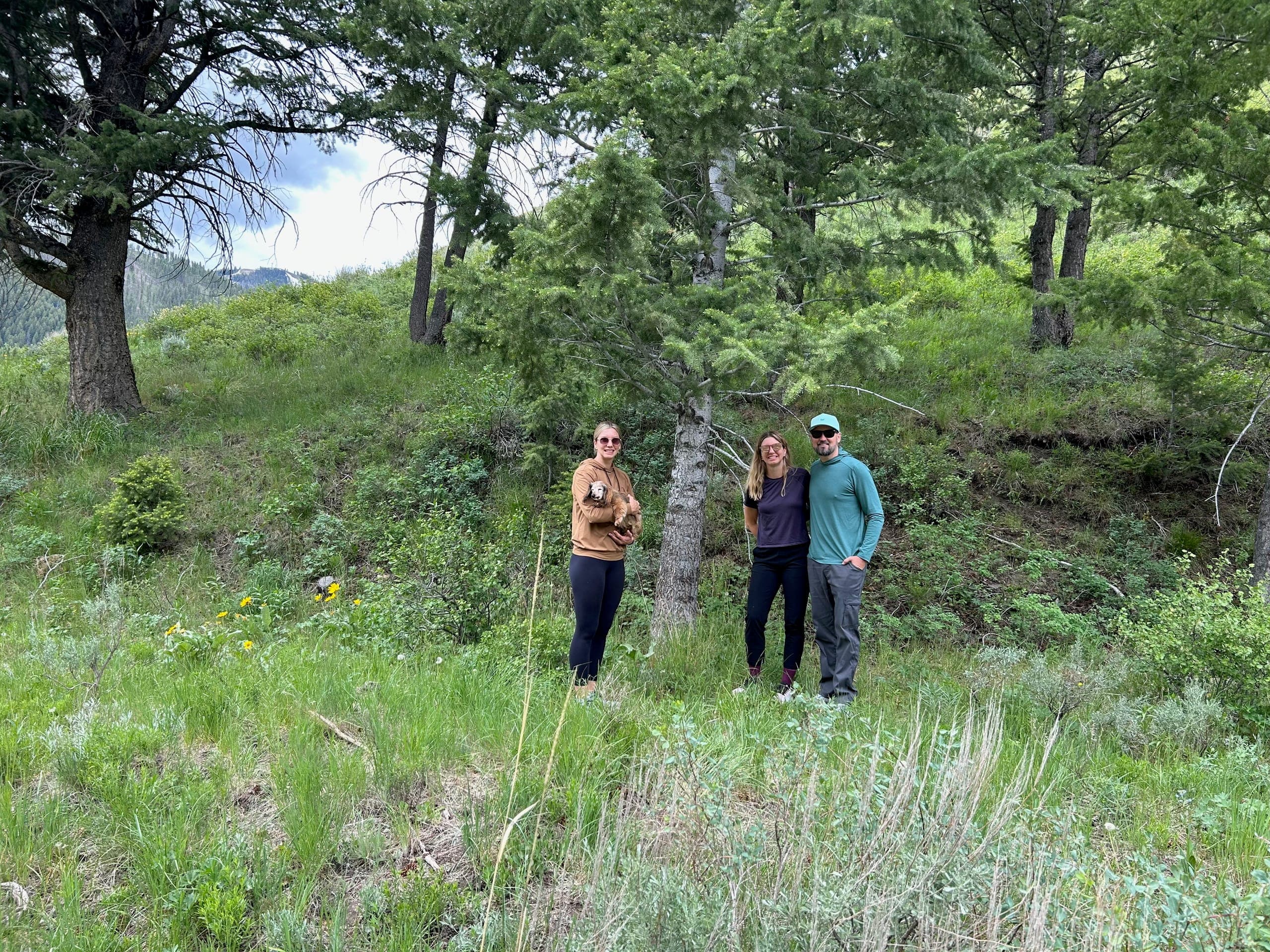 Group photo of Erik, Sarah, and Kayti holding Baby Dog in front of a tree on a green hillside.